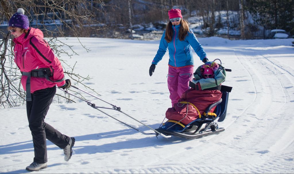 Pieni lapsi istuu Snow Comfort -kelkassa lämpöpussin sisällä, hänestä näkyy vain naama. Nainen työntää kelkkaa lumella ja toinen nainen vetää sitä vetoaisoista. On talvi ja aurinko paistaa.