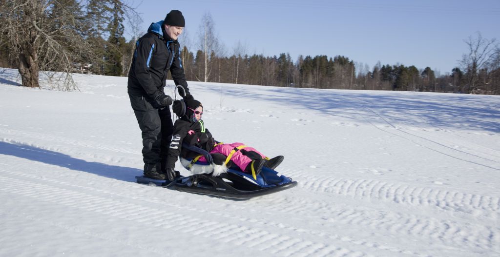 On aurinkoinen talvipäivä ja vaikesti vammainen tyttö laskee isänsä kanssa mäkeä Snow Comfort -kelkalla. Tytölle on kelkassa pään- ja vartalontuki. Isä seisoo kelkan takana olevalla jalustimella. Maisema on avara, luminen ja loiva mäki. Taustalla näkyy puita.