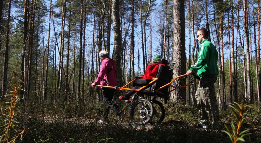 retkeilijä istuu Joelette-maastokärryssä. Toinen avustaja vetää kärryä avustusaisoista ja toinen työntää.