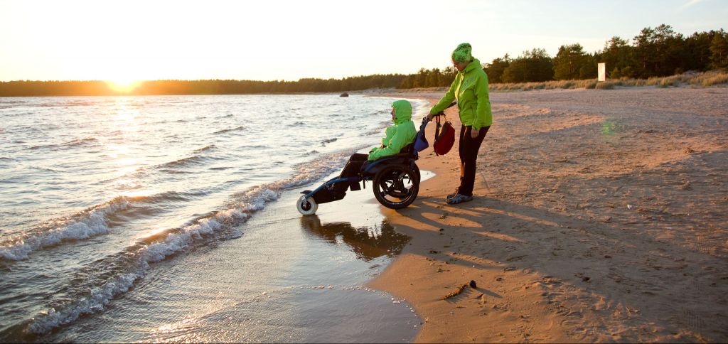 Kaksi henkilöä katselee auringonlaskua hiekkarannalla. Toinen heistä istuu Hippocampe-maastopyörätuolissa ja toinen työntää maastopyörätuolia. Molemmilla on keltavihreät takit päällään.