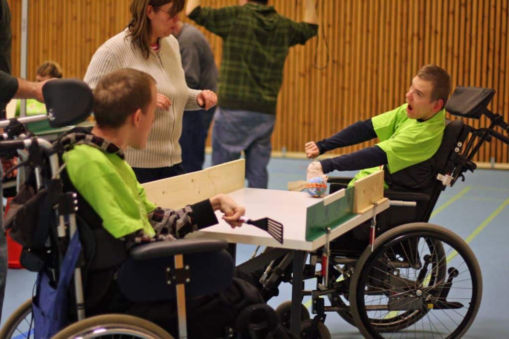 Kaksi nuorta miestä pelaa sovellettua pöytäpeliä. Molemmilla on paistinlasta pelimailana ja päällään kirkkaan vihreät Malike-paidat. Pöydän vieressä seisoo nainen, joka avustaa pelaamista.