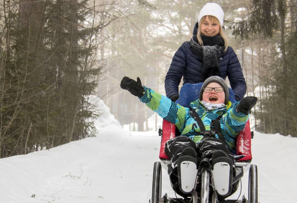 On talvi ja hieman sumuinen ilma. Kuvan oikeassa reunassa näkyy poika, joka istuu Kangoo Monitoimirattaissa kädet ylös kohotettuina ja nauravana. Nainen työntää rattaita. Kuvan vasemmassa reunassa näkyy lunta ja puita sumuharson takana.