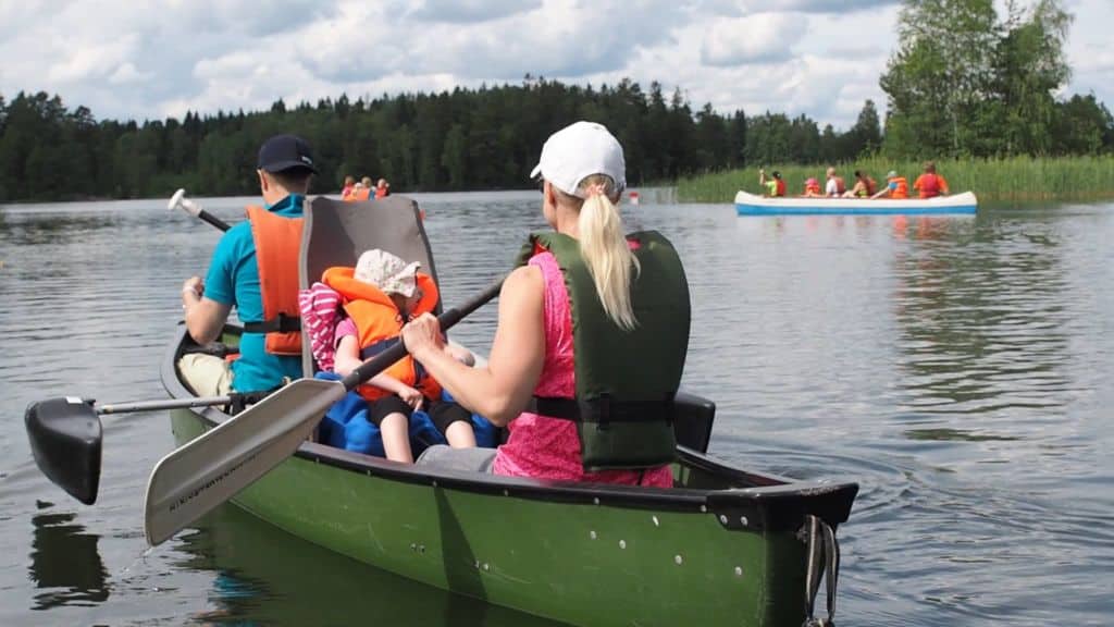 Kanootissa istuu nainen, lapsi ja mies. Lapsi istuu korkeaselkänojaisessa kanoottituolissa. Nainen ja mies melovat. Taustalla näkyy kaksi muuta kanoottia, joissa ihmisiä melomassa järvellä..