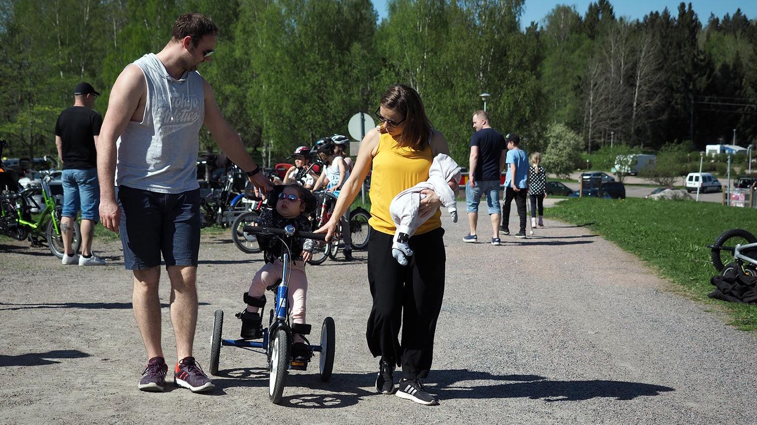 Perhe, jossa pieni lapsi pyöräilee ja vanhemmat tukevat. Äidin sylissä pieni vauva.