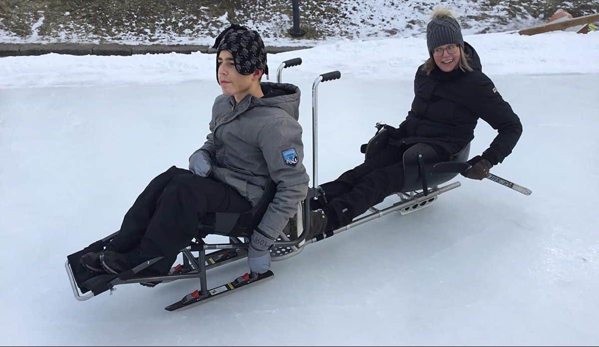 Nuori mies istuu jäällä luistelukelkassa ja hänen takanaan istuu nainen jääkiekkokelkassa. Naisella on käsissään lyhyet kelkkakiekkoon sopivat jääkiekkomailat.