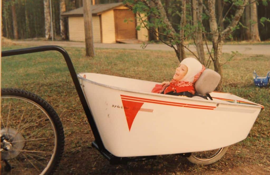 Vanha valkoinen polkupyörän peräkärry Malikkeen alkuajoilta. Peräkärryssä istuu nauravainen tyttö, taustalla nurmikenttää ja lautarakenteinen beige autotalli.