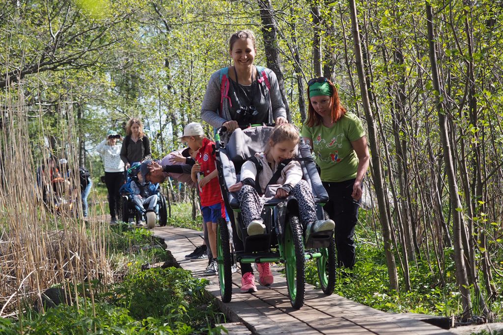 Ihmisiä kulkee jonossa pitkospuita pitkin. Kolme lasta istuu maastovälineissä ja aikuiset työntävät välineitä.