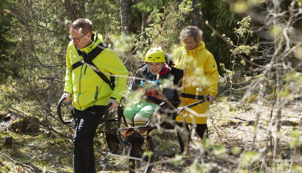 Poika istuu Joelette maastokärryssä, jota mies vetää ja nainen työntää polkua pitkin metsässä. Pojalla on päässään keltainen kypärä. Naisella ja miehellä on yllään keltaiset takit.