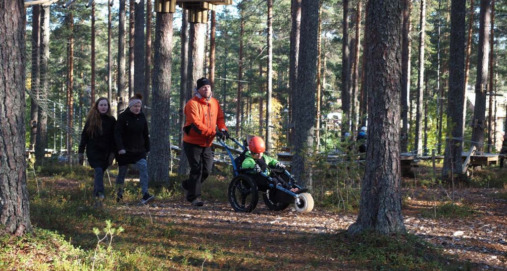 Vihreä takkinen poika istuu Hippocampe maastopyörätuolissa ja oranssi takkinen mies työntää maastopyörätuolia polkua pitkin mäntymetsässä.. Kaksi naista kävelee heidän perässään. Naisilla on mustat takit. On syksy, mutta aurinko paistaa ihanasti.