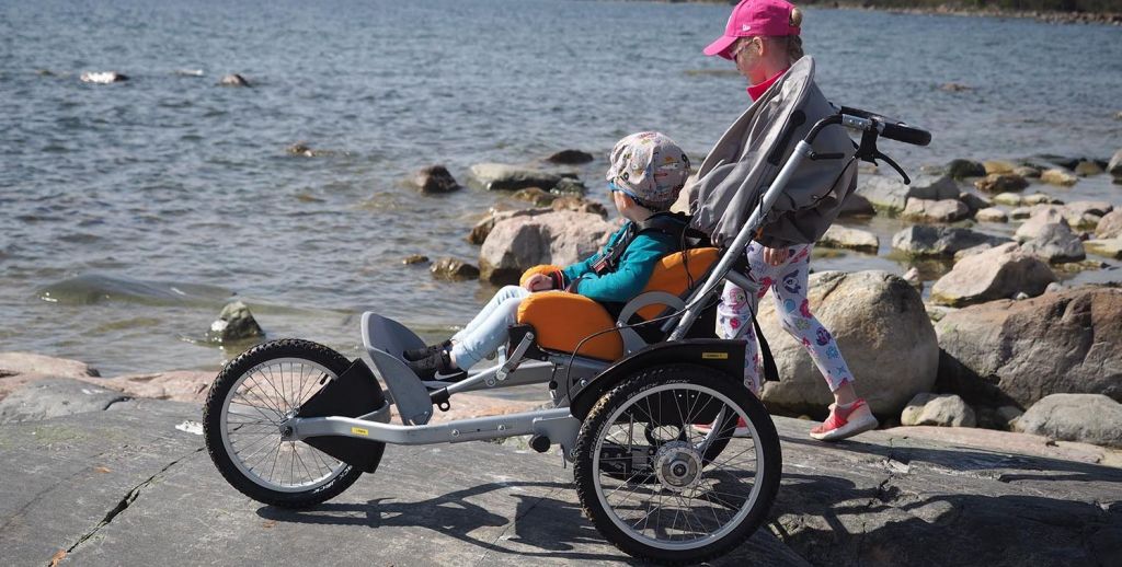 Lapsi istuu Kimba-rattaissa kalliolla meren rannalla. Toinen lapsi seisoo rattaiden takana. On kesä ja aurinko paistaa.