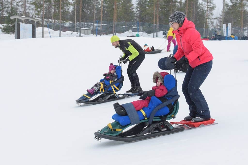 On talvi, kaksi lasta istuu Snow Comfort -kelkoissa ja kaksi aikuista seisoo kelkkojen takana kyydissä. He laskevat vierekkäin alas loivaa mäkeä.