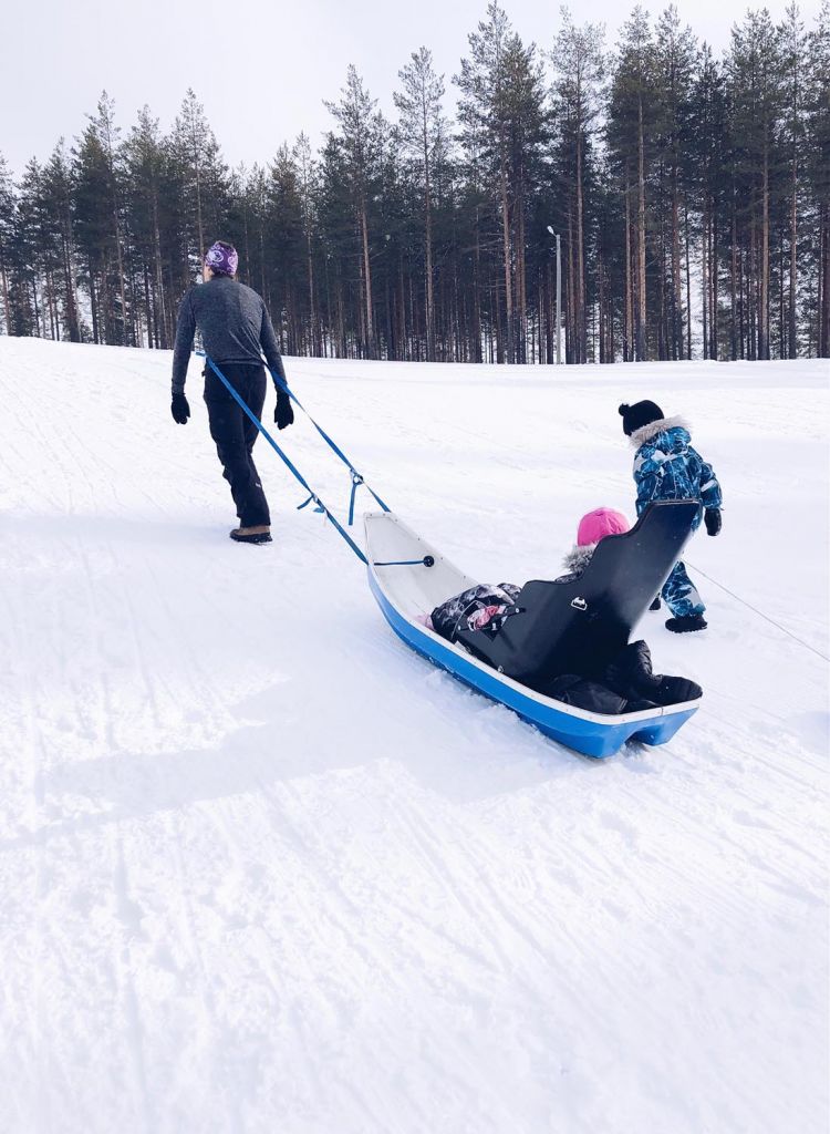 Tyttö istuu ahkiossa, jossa on istuin. Hänen isänsä vetää ahkiota aisoilla ja hänen veljensä kävelee vieressä.