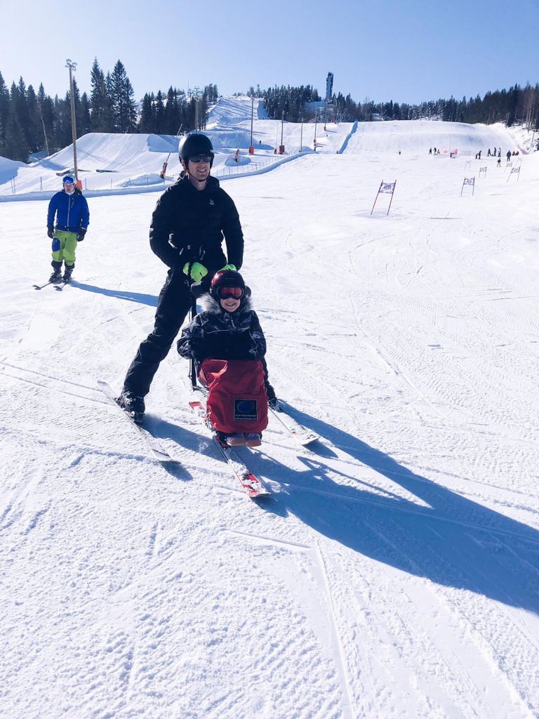 Tyttö istuu laskettelukelkassa ja isä ohjaa kelkkaa. Molemmat hymyilevät.