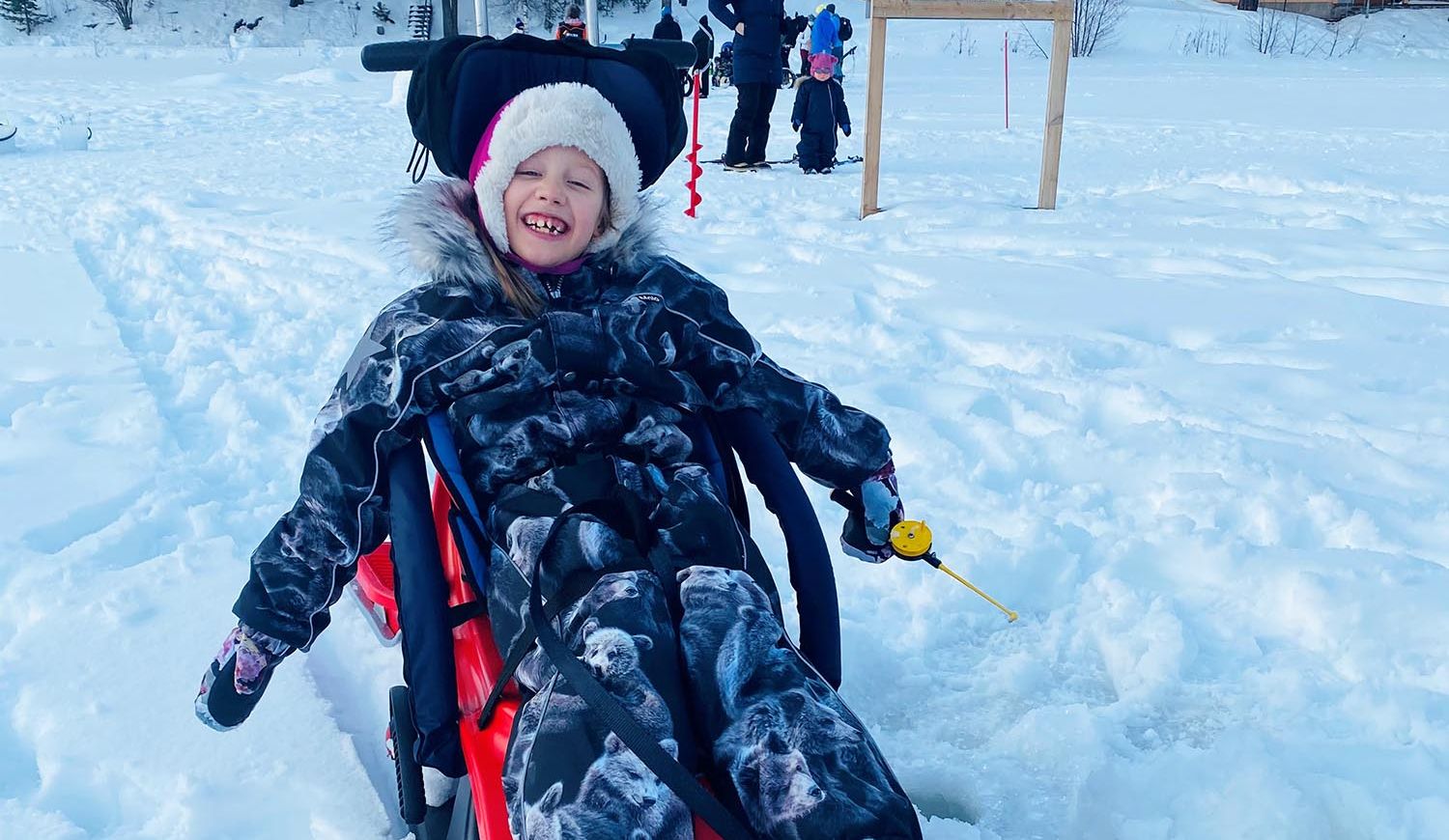 Vaikeasti vammainen tyttö istuu Snow Comfort -kelkassa, jossa on tukeva selkäosa. Hänen kädessään on pilkki.