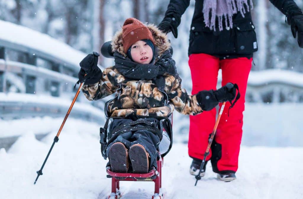Poika hiihtää hiihto- ja luistelukelkalla istuen. Kelkassa on selkänoja ja niskatuki, jotka mahdollistavat vaikeasti vammaisen lapsen hyvän asennon kelkassa.