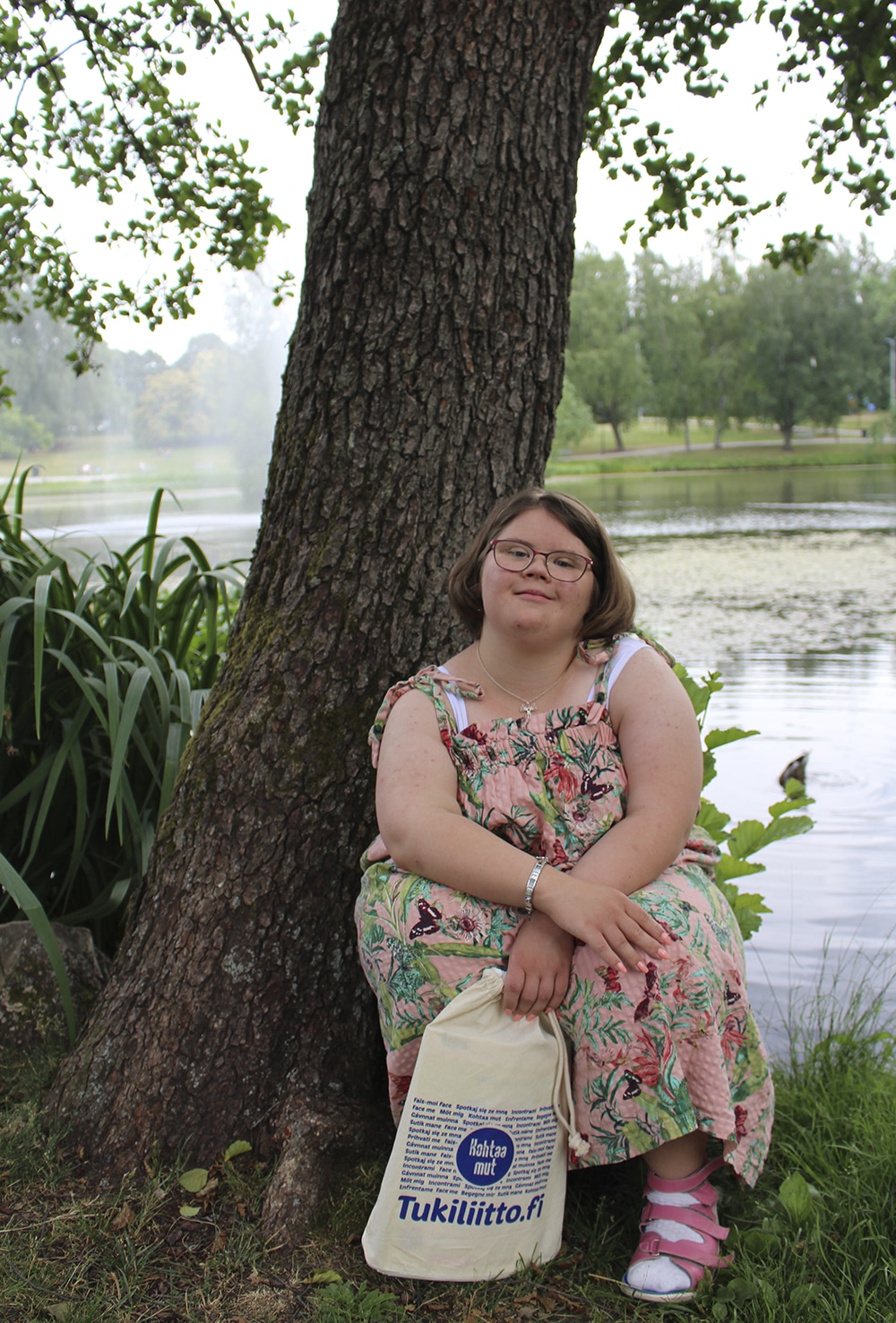Silja Nuottimäki istuu puun edessä kesäpäivänä. Päällä hänellä on kesämekko ja kädessä hän pitää Tukiliiton pussukkaa. 