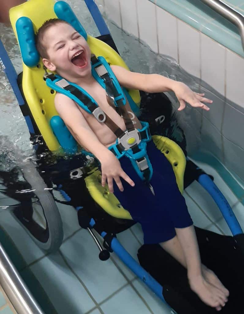 Lapsi nauraa uima-altaassa. Keltainen kylpytuki on sijoitettu rattaisiin uimisen ajaksi.