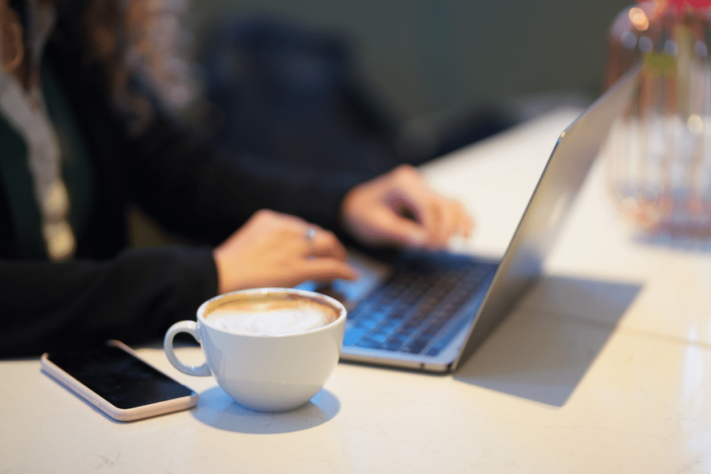 Pöydällä on sivusta kuvattuna kannettava tietokone ja sen näppäimistöllä ihmisen kädet. Tietokoneen vieressä on kahvikuppi.