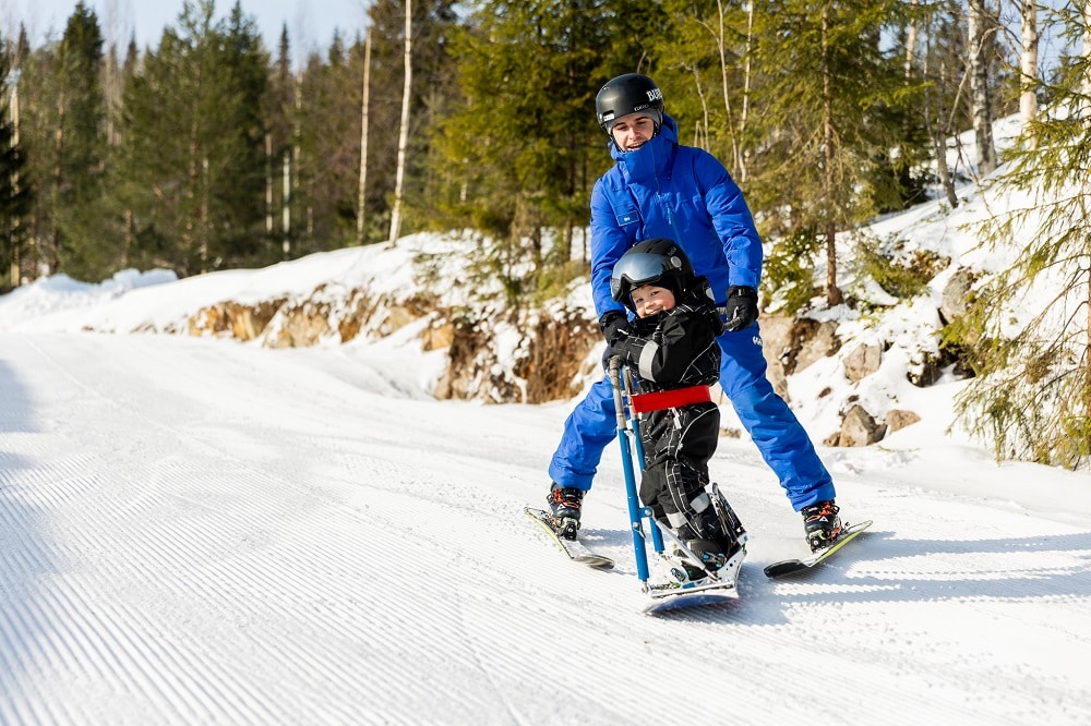 Poika laskee avustajan kanssa lumilaudalla mäkeä.