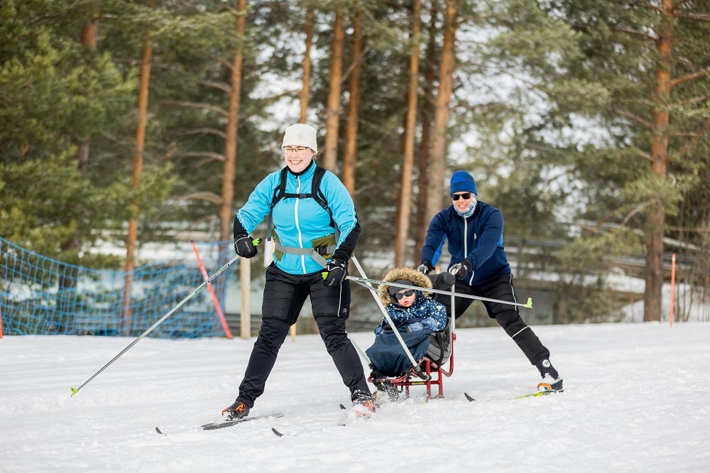 Perhe hiihtää. Äiti vetää hiihtokelkkaa, lapsi istuu kyydissä ja isä kelkan takana. Kaikki hymyilevät iloisesti.