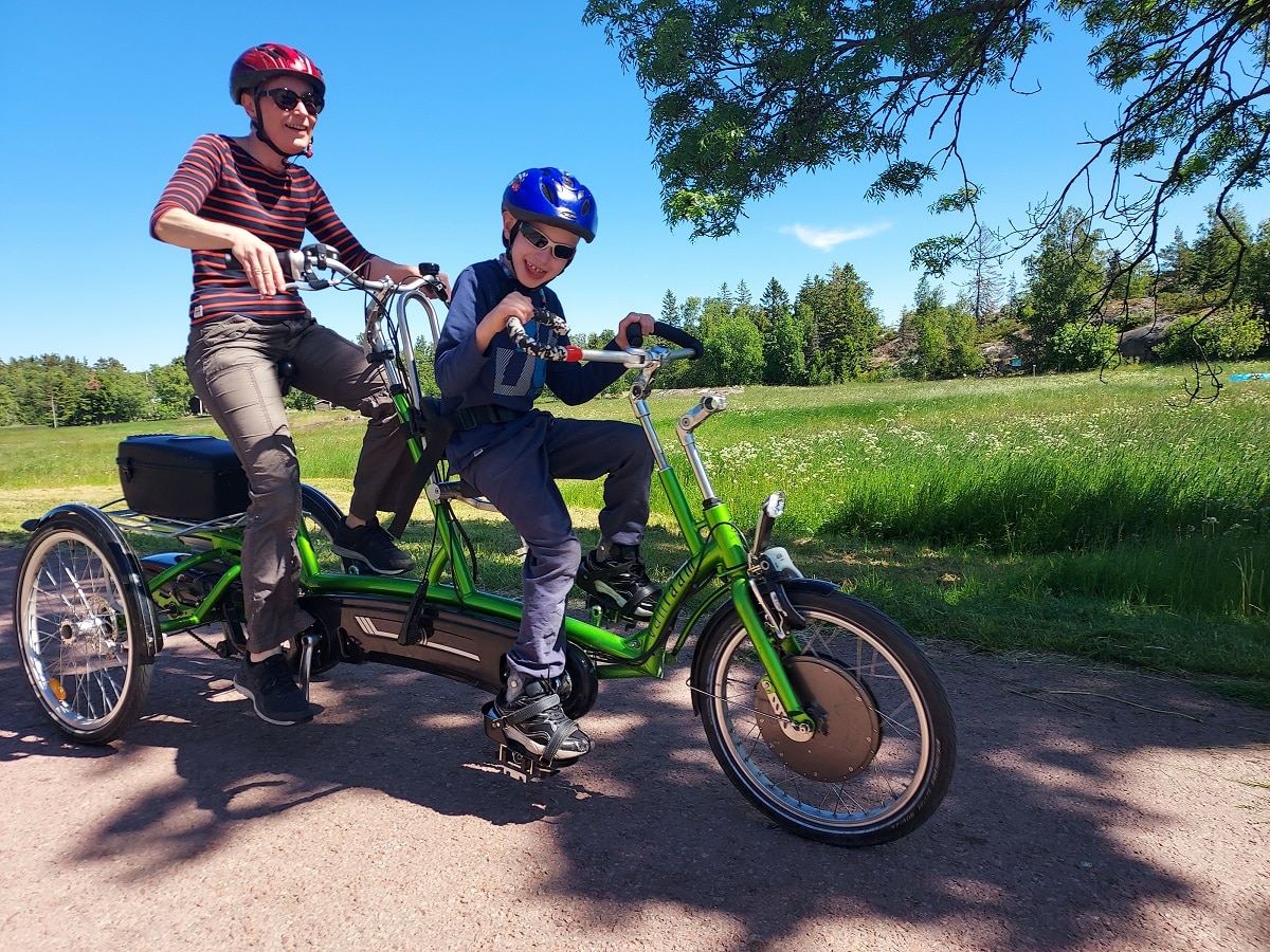 Lapsi ja aikuinen ajavat vihreällä tandem-pyörällä Ahvenanmaalla. On kesä ja kummallakin on aurinkolasit ja he hymyilevät. Erityispyörät mahdollistavat pyöräilyä kaikille.
