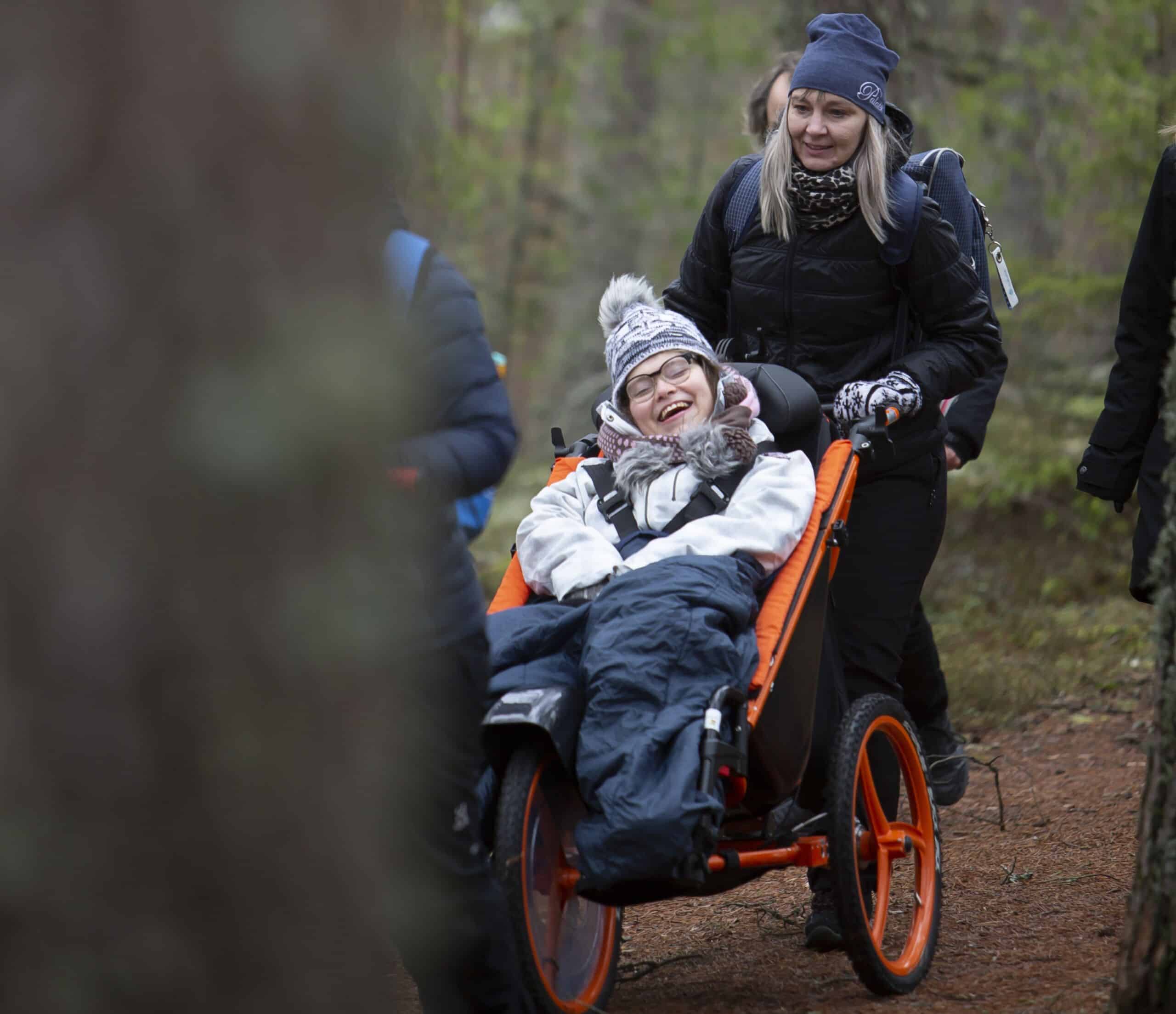 Nainen valkoisessa takissa harmaa pipo päästää hymyilee ja nauttii metsäretkestä. Mustatakkinen nainen työntää häntä maastopyörätuolissa metsäpolulla.