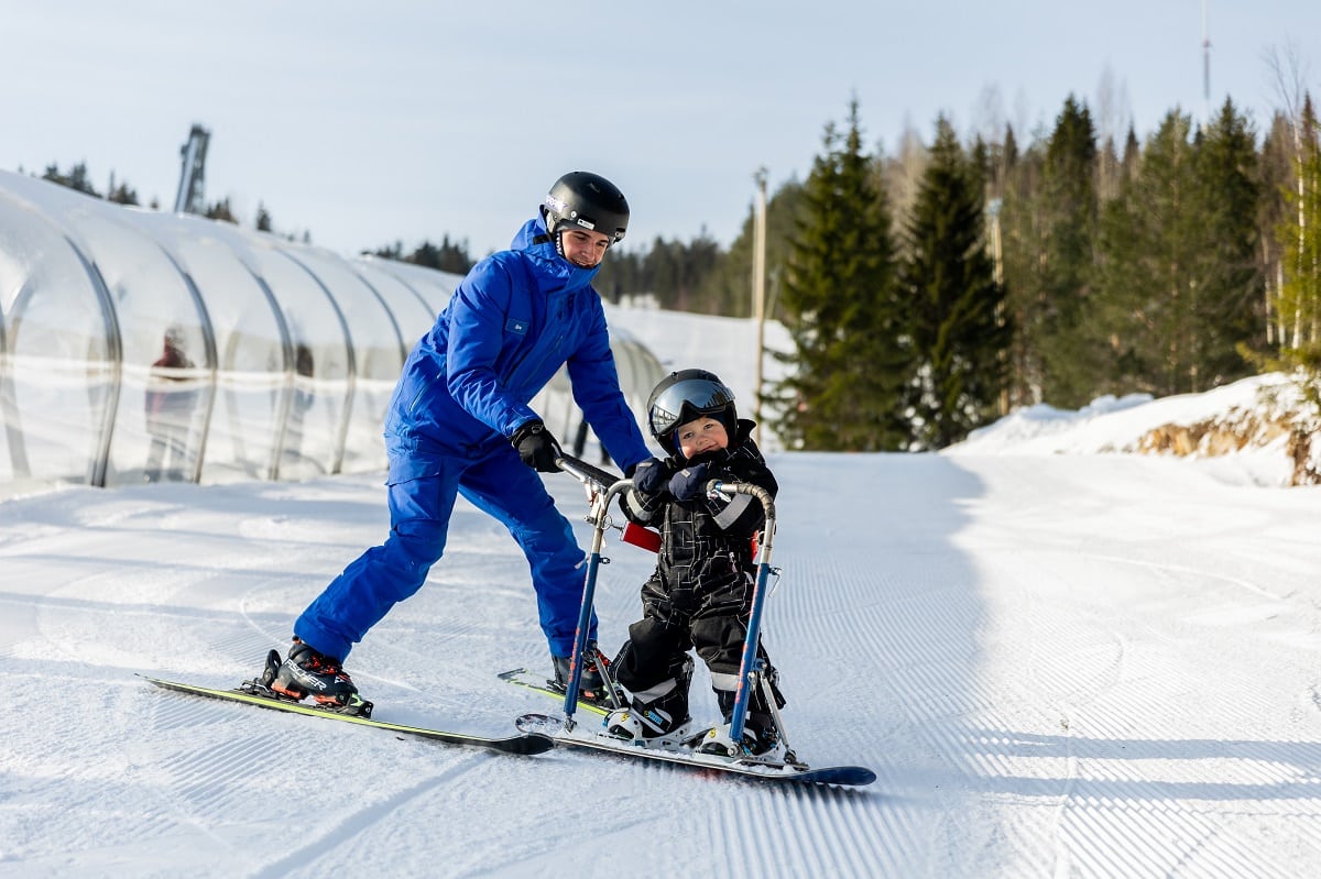 sinipukuinen mies laskettaa lumilaudalla olevaa pikkupoikaa rinnettä alas. Pojalla on musta haalari ja hän hymyilee iloisesti.