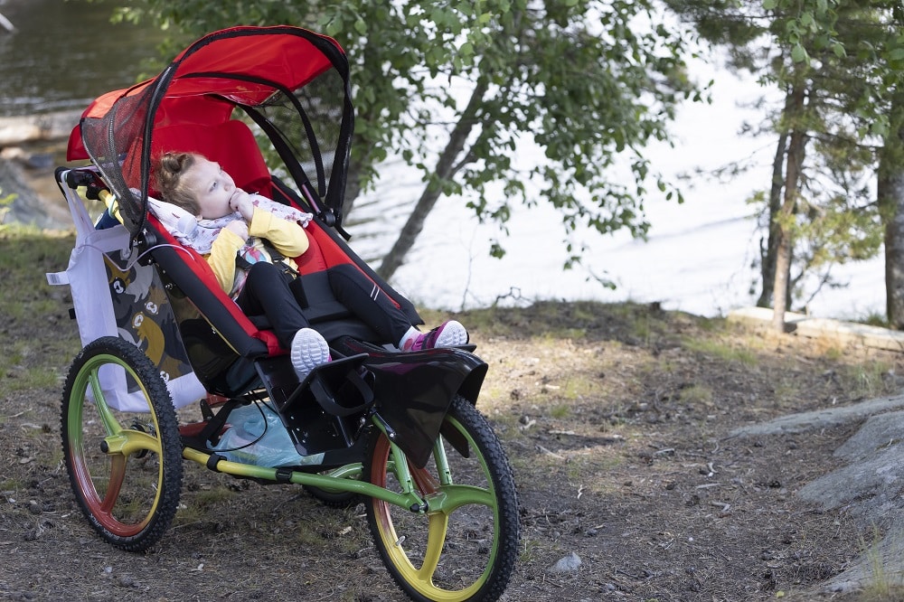 Isorenkainen monitoimirattaat lapselle, joka istuu nauttimassa järvimaisemista kesällä rattaiden punainen aurinkosuoja kohotettuna.
