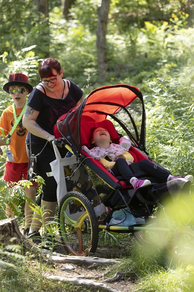 Perhe ulkoilee kesäisessä metsässä. Äiti työntää punakuomuisissa rattaista nauravaa lasta. Aurinkolasipäinen poika hellehatussaan kurkistelee äidin takaa.