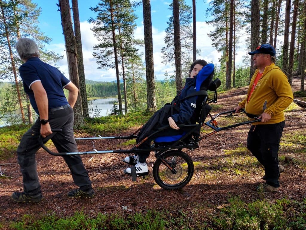 Sinipaitainen mies vetää kaksipyöräistä maastokärryä jossa istuu iloinen retkeläinen. Keltatakkinen mies lippalakissa työntää kärryä sen takaa hymyillen.