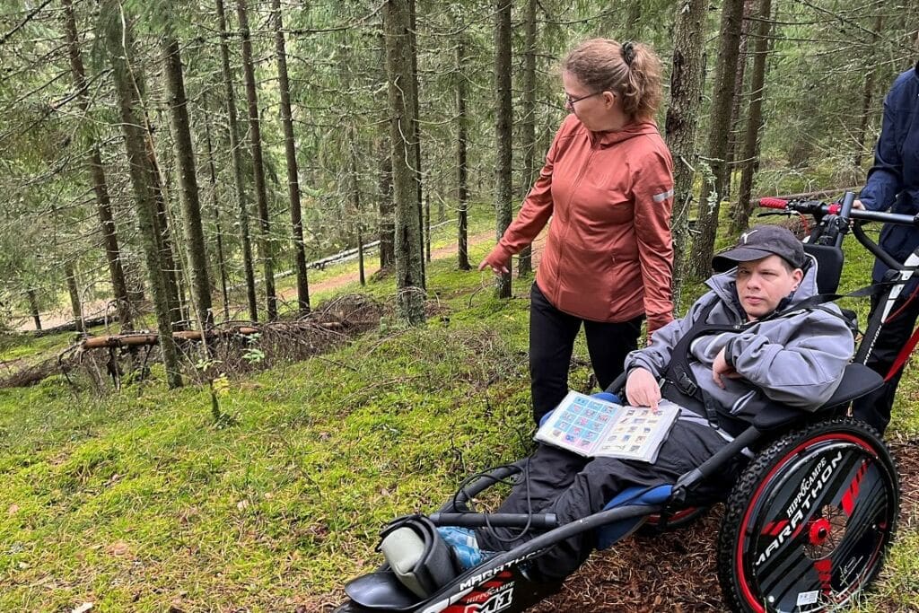 Mies istuu metsässä Maastopyörätuoli Hippocampen kyydissä ja hänellä on sylissään kuvia kommunikointiin. Käsi osoittaa yhtä kuvaa. Miehen rinnalla seisoo nainen pukeutuneena ruosteenpunaiseen ulkoilutakiin ja mustiin housuihin. Nainen katsoo miehestä pois päin alas viettävään rinteeseen. Maastopyörätuolin työntöaisalla näkyy sitä työntävän, ihmisen kädet.