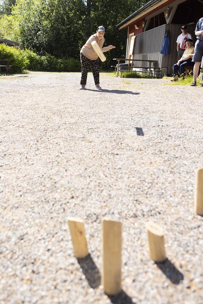 Vanhempi nainen heittää mölkkyä kuvan taustalla. Kuvan edessä on mölkkypalikoita. Kuvan sivussa on kolme ihmistä katselelmassa mölkynheittoa.