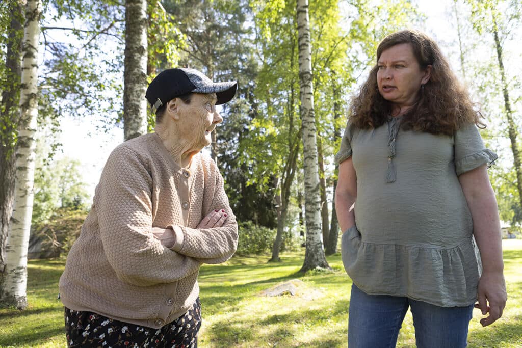 Kuvassa kaksi naista keskustelevat keskenään. Vasemmalla on vanha nainen kädet puuskassa ja oikealla keski-ikäinen nainen kädet housuntaskuissa sanomassa jotakin.