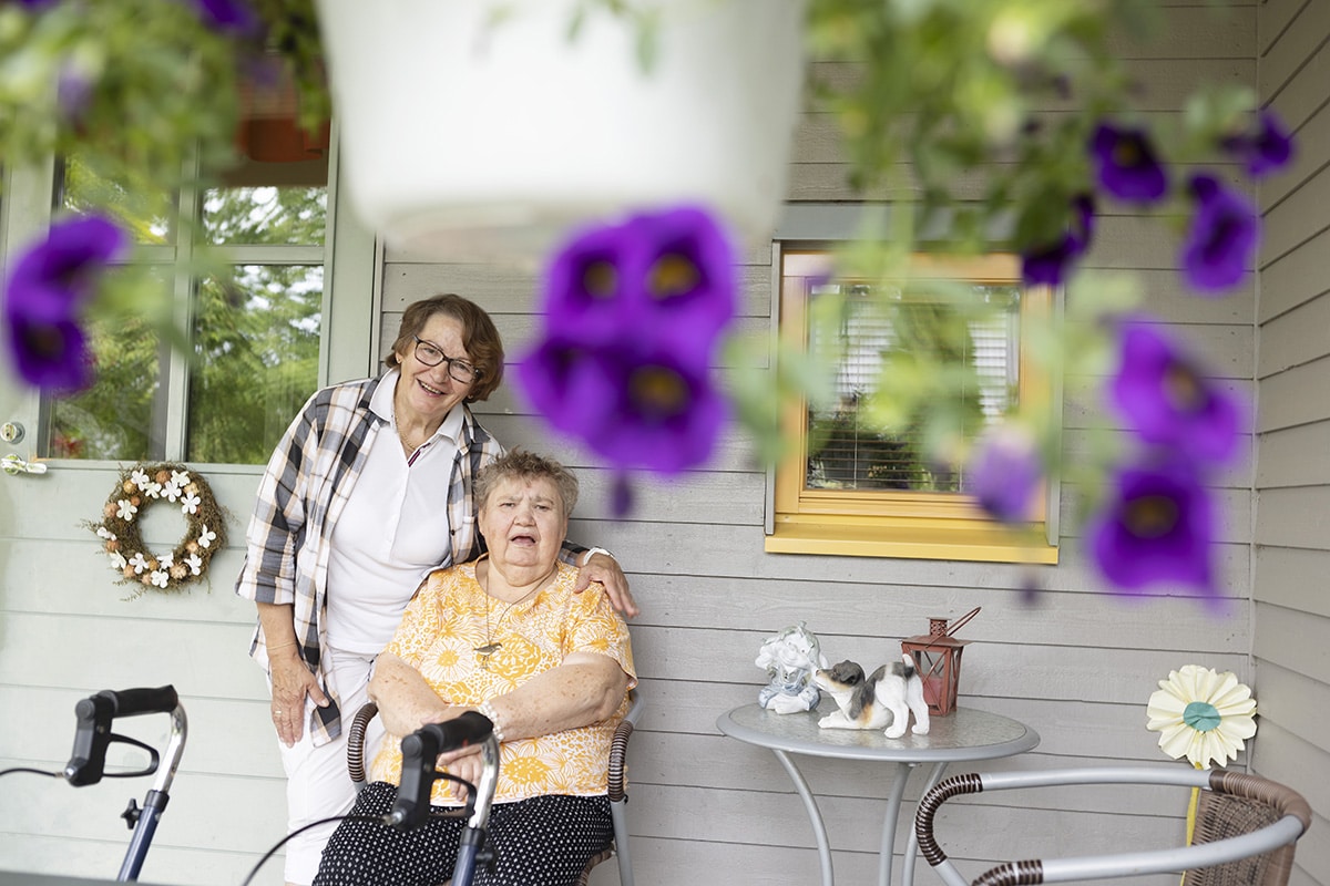 Kuvassa on etualalla violetti riippuva kesäkukka. Takana kaksi naista hymyilevät talon seinän edessä. Toinen nainen istuu tuolissa ja toinen on hänen takanaan pitäen toisen naisen olkapäällä kättä.
