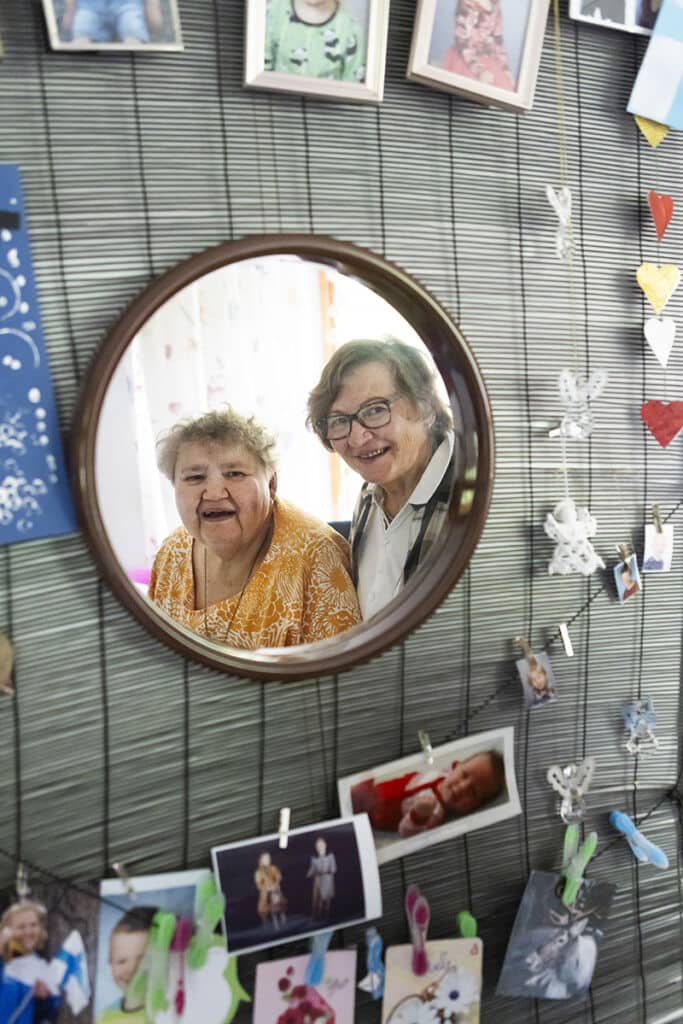 Kuvassa on keskellä peili, josta näkyy kahden nauravaisen vanhemman naisen kasvot. Peilin ympärillä on seinässä valokuvia kiinnitettynä pyykkipojilla naruun.