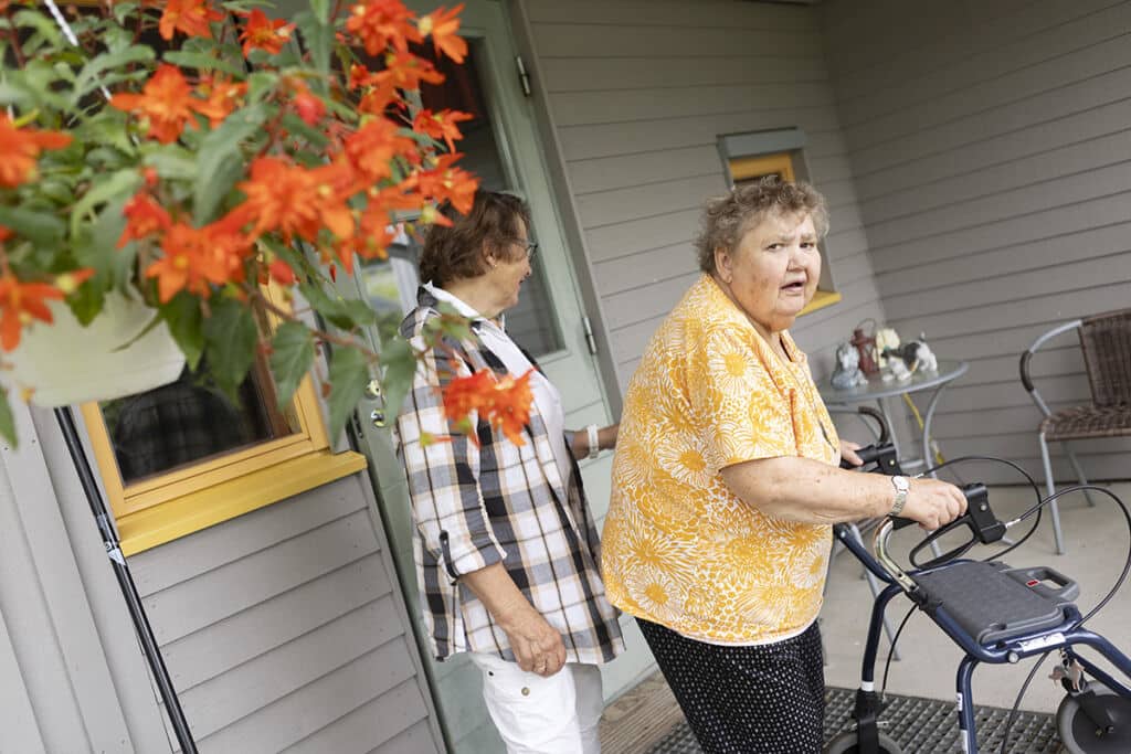 Kuvan etualalla on oranssi katosta riippuva kesäkukka. Kesäkukan takana talon edustalla seisoo kaksi vanhempaa naista. Heistä toinen katsoo kameraan ja kulkee rollaattorilla.