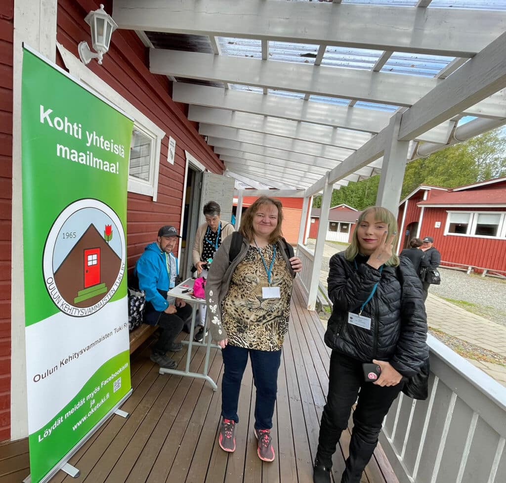 Kaksi naista hymyilee kameralle. He seisovat kyltin vieressä, jossa lukee "Kohti yhteistä maailmaa". Heidän takanaan on muitakin ihmisiä. He ovat kaikki punaisen talon terassilla.