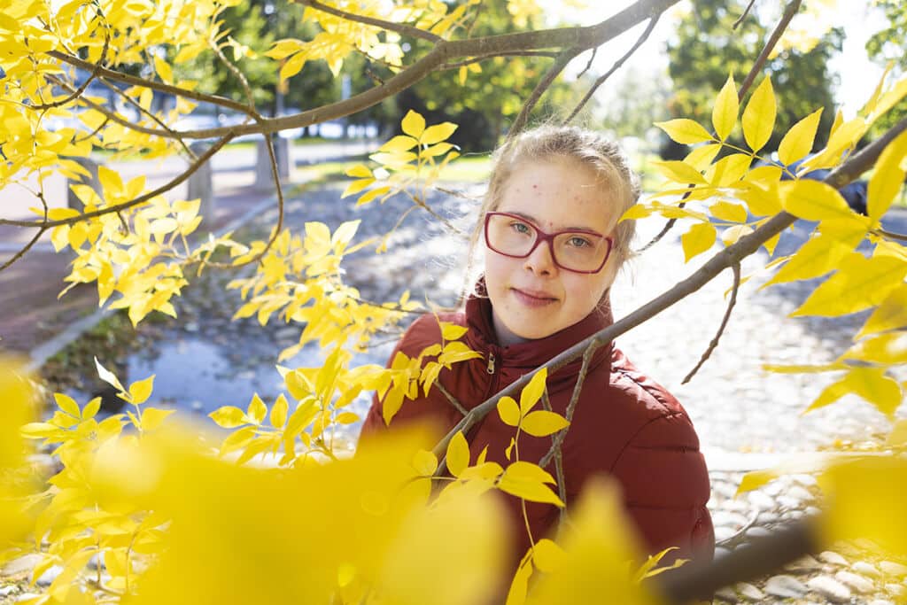 Anniina Hirvonen päättää asioista omassa kodissaan. Kuvassa Anniina seisoo puiden oksien takana. Oksissa on paljon keltaisia lehtiä.