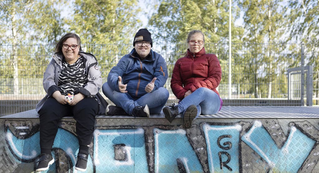 Jere Hirvonen, Ann-Mari Saastamoinen ja Anniina Hirvonen asuvat Joensuussa. Kuvassa he istuvat vierekkäin penkillä ja hymyilevät kameralle.