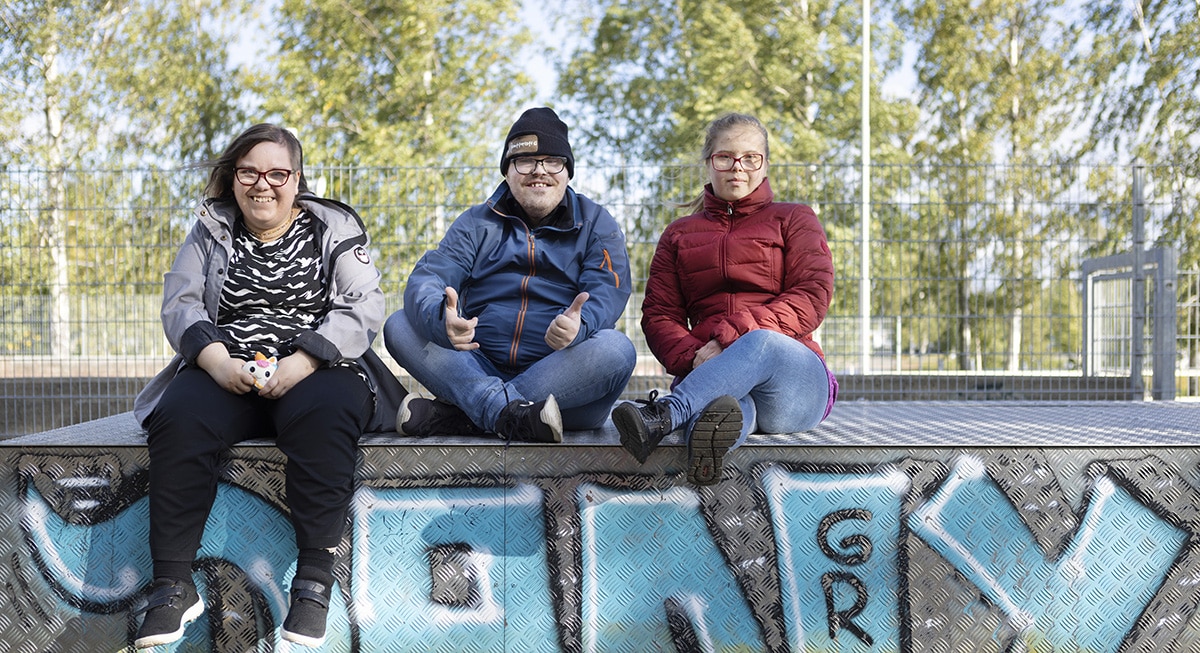 kolme ihmistä istuu graffiti-aidalla ja hymyilee