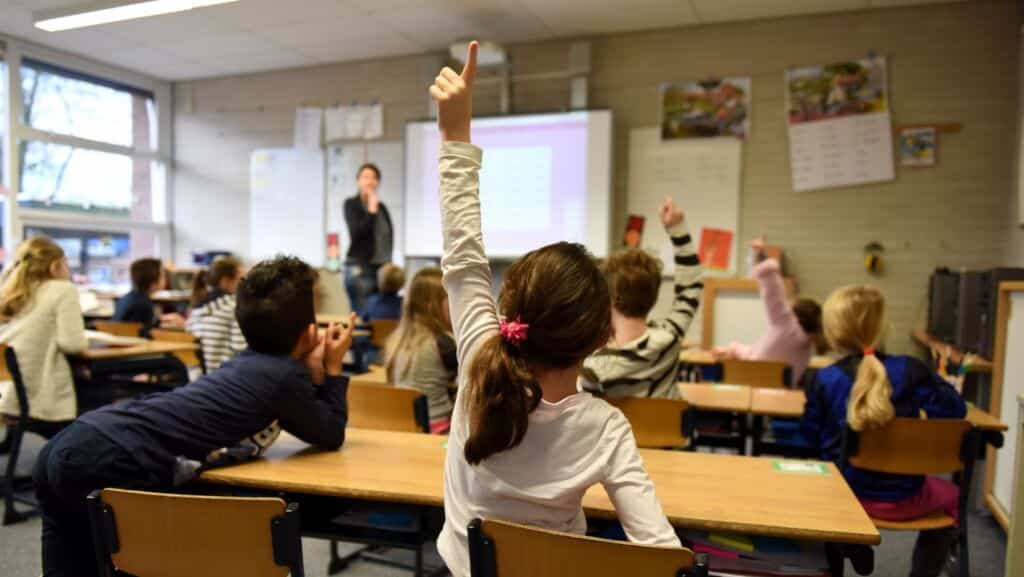 Oppilaat ovat koululuokassa pulpettien ääressä ja edessä on opettaja. Yksi oppilaista viittaa.
