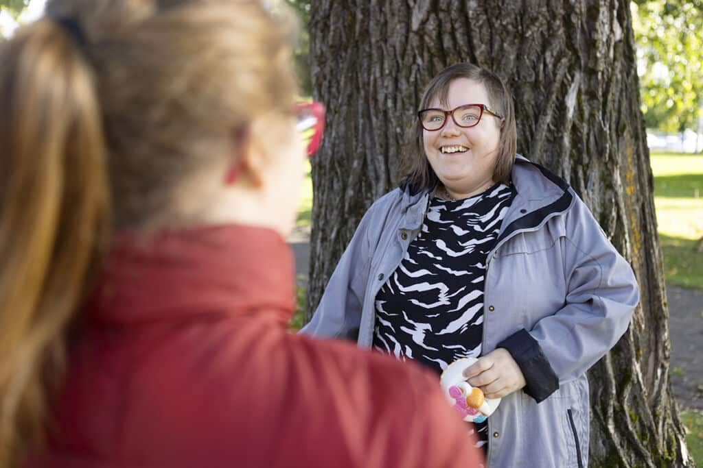 Ann-Mari Saastamoinen harjoittelee raha-asioiden hoitamista ja kelloa. Kuvassa hän seisoo puun edessä ja hymyilee Anniina Hirvoselle.