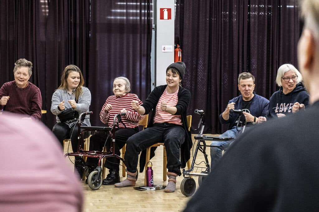 Teatterin ohjaaja istuu tuolilla keskellä ja muut ihmiset istuvat hänen ympärillään kuunnellen.