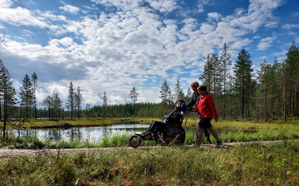 Luontomaisema, jossa mies ja nainen vaeltavat työntäen maastorattaissa istuvaa nuorta. Yhdenvertaisesti luontoon -retkellä Pudasjärvellä.