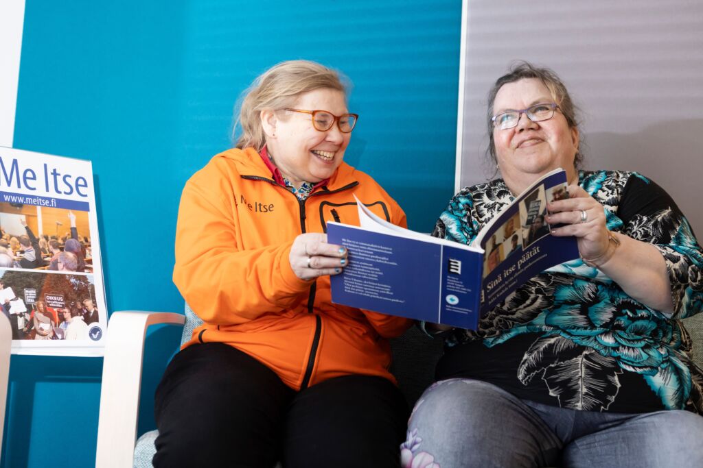 Kaksi naista lukevat kirjaa sohvalla. Heidän vieressään on MeItse-kyltti.