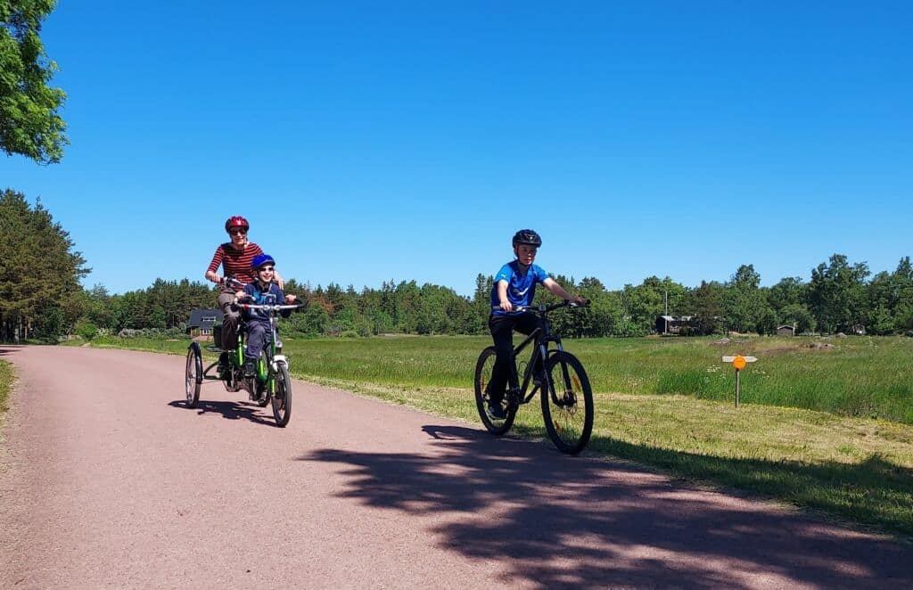 On kesä. aurinko paistaa. Aikuinen ja lapsi pyöräilevät tandempyörällä. Toinen lapsi pyöräilee vieressä yksin.