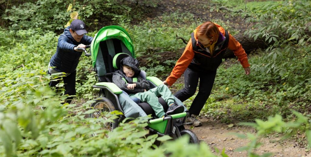 Kaksi aikuista ja yksi lapsi nousevat ylämäkeen kapella polulla. Lapsi istuu vihreissä maastorattaissa. Toinen aikuinen työntää rattaita takaa ja toinen vetää sivulta. Ympäristö on vehreä. Sää on harmaa. Kaikki henkilöt ovat pukeutuneet välikauden ulkoiluvaatteisiin.