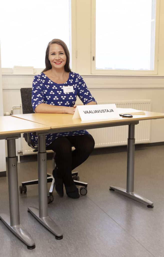 Nainen istuu pöydän takana ja hymyilee kameralle. Hänen edessään pöydällä on kyltti, jossa lukee "vaaliavustaja".