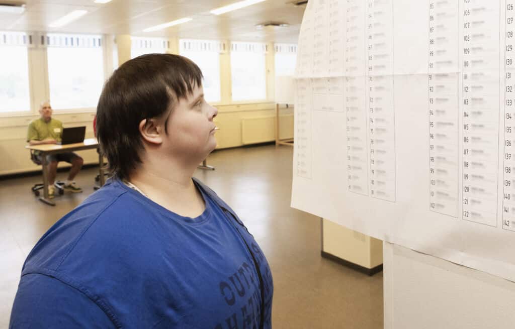 Nainen katselee julistetta, jossa on näkyvillä äänestysnumerot. Hänen takanaan on äänestyspöytä, jonka ääressä istuu ihminen.