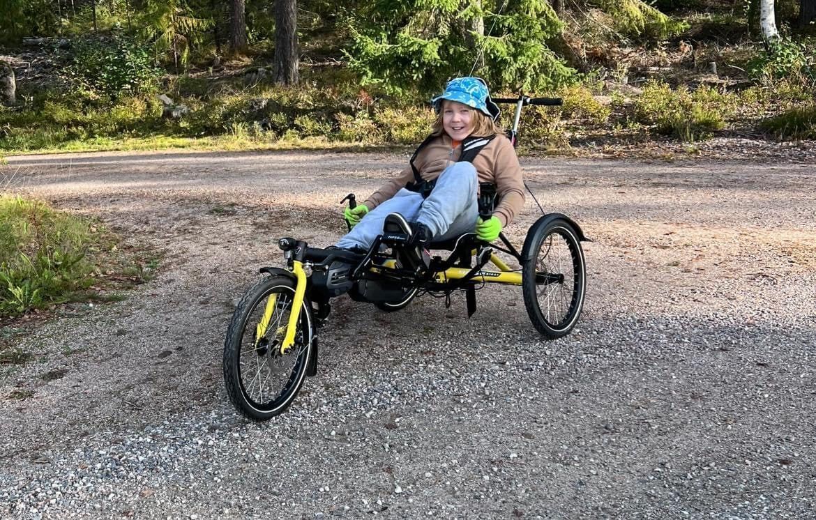 Poika istuu nojapyörässä hiekkatiellä. Nojapyörässä on istuin, jossa kuljettaja on puolimakaavassa asennossa. Nojapyörässä on kolme rengasta, kaksi takana ja yksi edessä. Polkimet ovat eturenkaan lähellä niin, että polkijan jalat osoittavat eteenpäin.